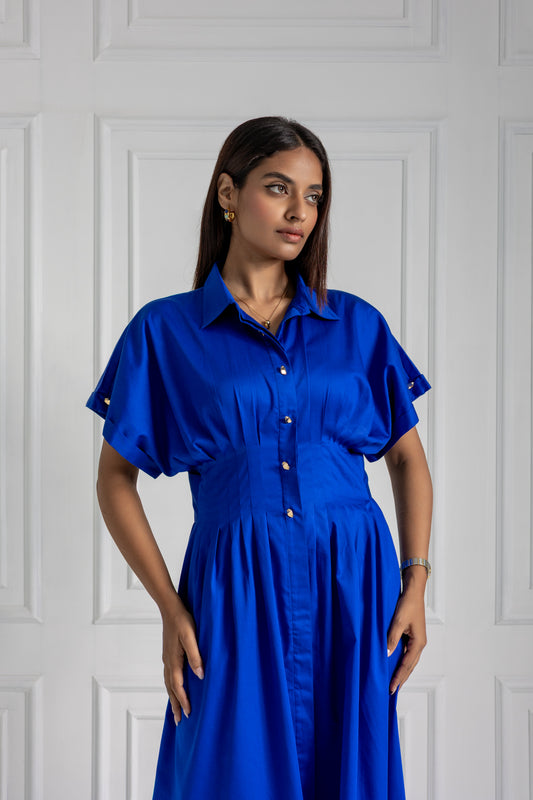 Woman wearing a blue dress pleated at the waist against a white paneled wall